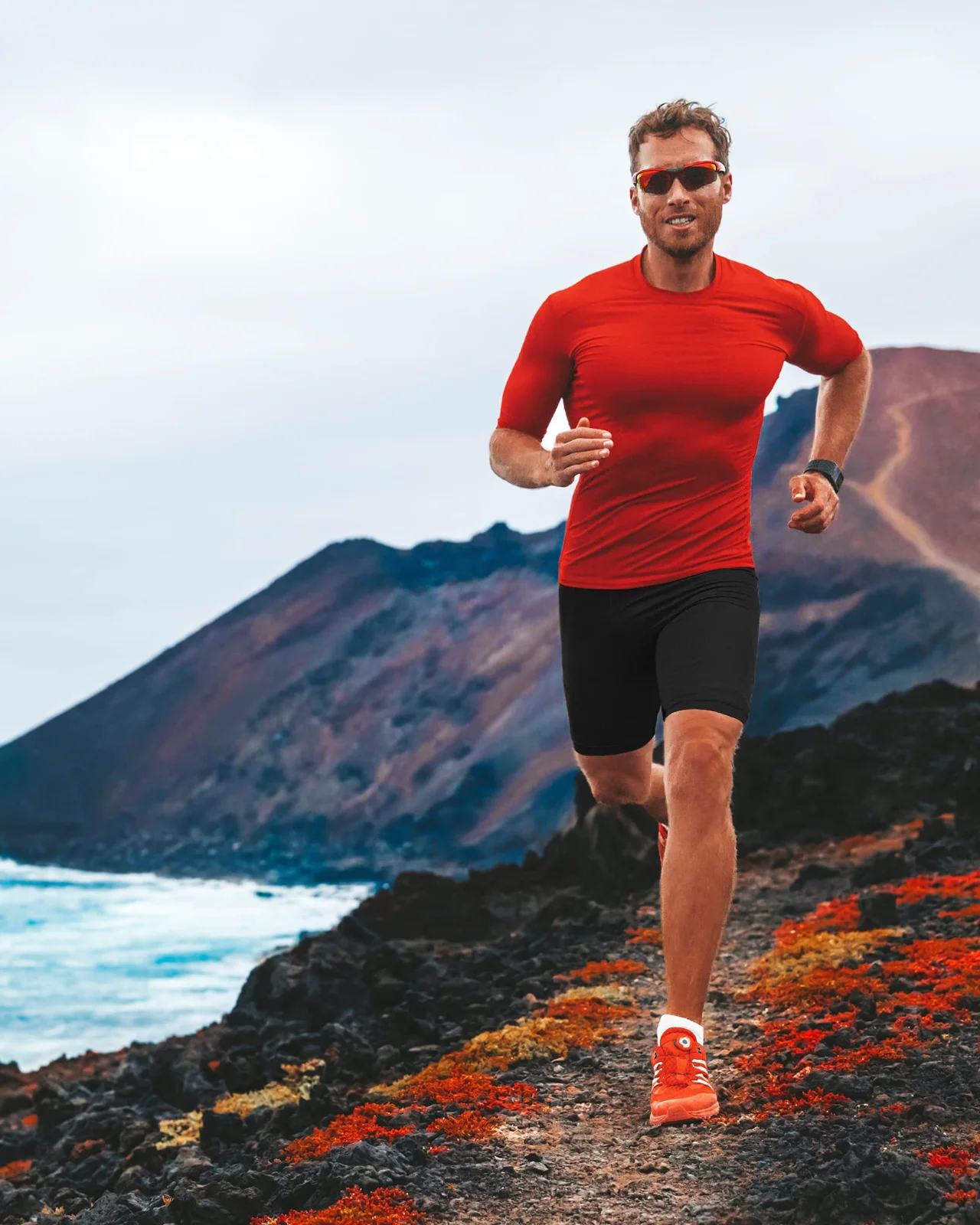 Black compression shorts under a red training tee on a coastal mountain road, mid-stride with the sea below