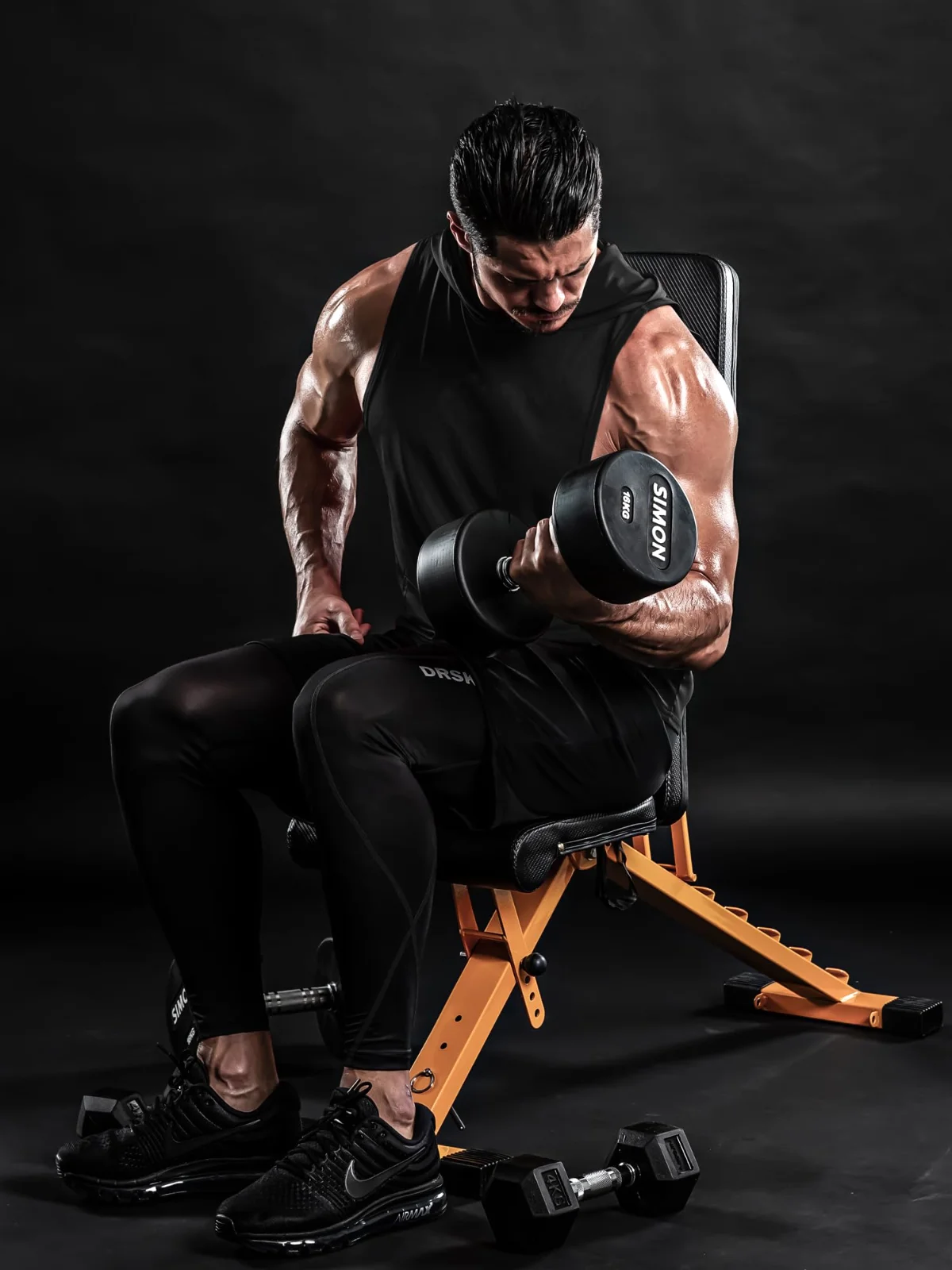 The black hooded tank seated on a bench, hood up, a dumbbell held at the shoulder
