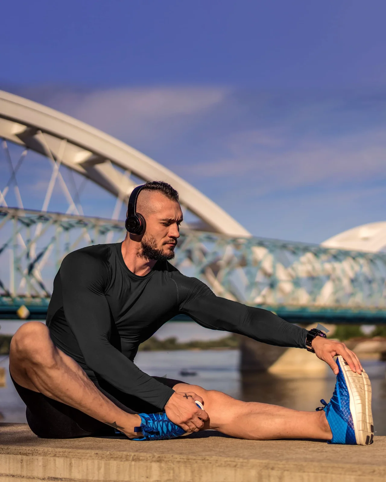 A black long-sleeve base layer stretching on a boardwalk under a steel bridge, one leg out straight